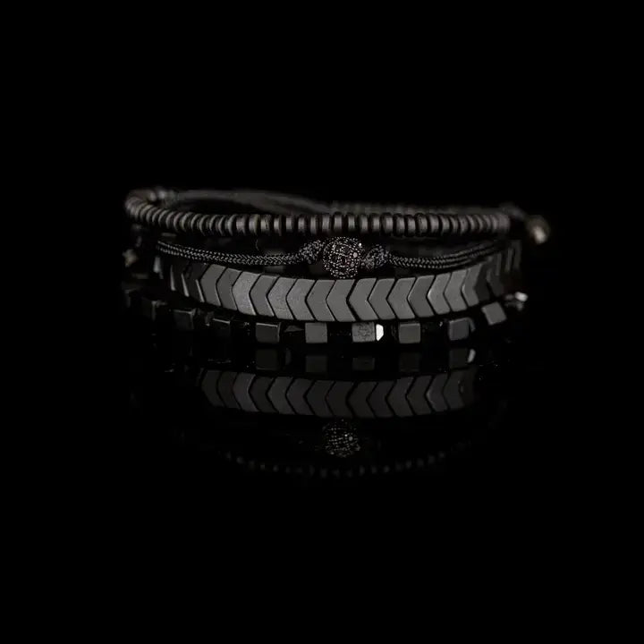Men’s layered bracelet set featuring gunmetal chevron metal beads, black hematite cube beads, dark metallic heishi beads, black braided cord, and a pavé-style gunmetal accent bead, stacked in a sleek monochromatic luxury design on a white background.