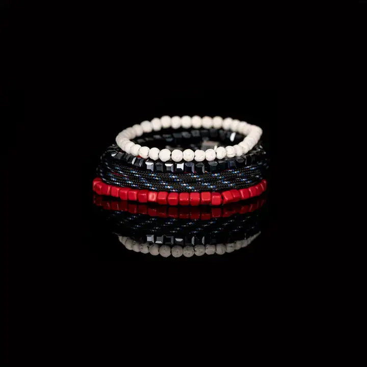 Men’s layered bracelet set featuring vibrant red cube beads, black braided cord with blue accents, faceted black hematite beads, and white heishi beads, stacked in a bold contemporary luxury design on a white background.