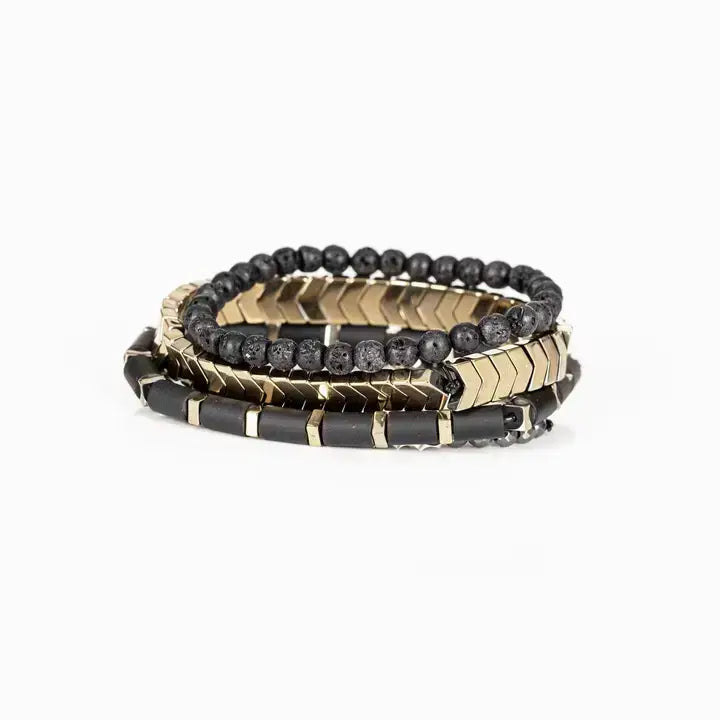 Men’s layered bracelet set featuring matte black lava stone beads, gold-tone chevron metal beads, black cylindrical beads with gold spacers, and faceted hematite beads, stacked in a modern luxury design on a white background.