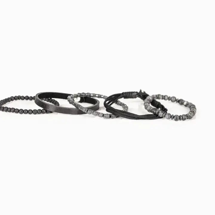Men’s layered bracelet set featuring gray marbled natural stone beads, a black leather cuff, matte black lava stone beads, black braided cord, hematite cube beads, and metallic spacer accents, stacked in a sophisticated monochromatic luxury design on a white background.