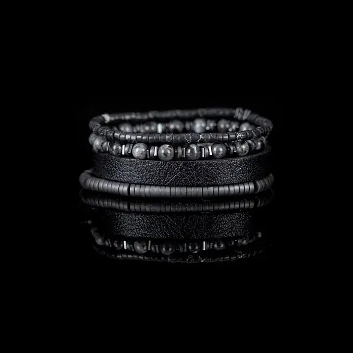 Black multi-layer men’s bracelet featuring textured leather band, gunmetal hematite bead strands, matte grey stone beads, and layered metallic accents on a light background.