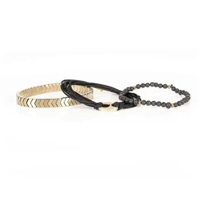 Black and gold layered men’s bracelet with lava stone beads, braided rope wraps, gold chevron metal bar, and metallic accents on white background.