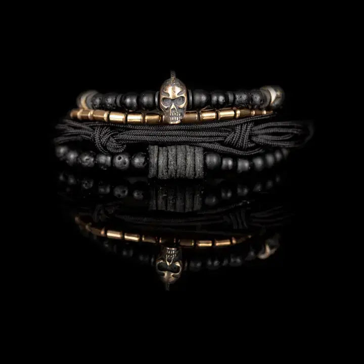 Black layered men’s bracelet set with lava stone beads, metallic skull centerpiece, gold-tone spacer accents, matte black beads, and braided rope wraps on white background.