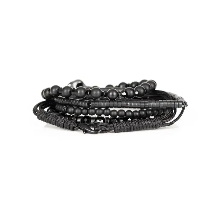 Black layered men’s bracelet set with matte black beads, silver skull centerpiece, faceted crystal accents, and braided rope wraps on white background.