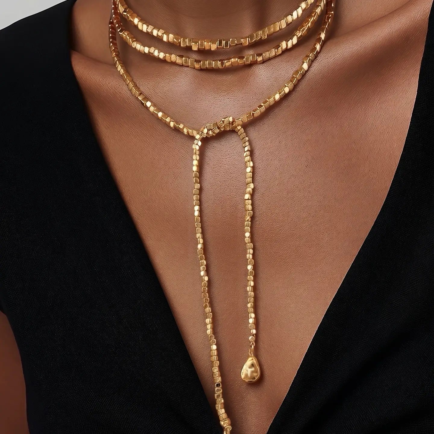 Layered gold necklace with three strands and long lariat drop chains ending in sculpted gold pendants on a white background.