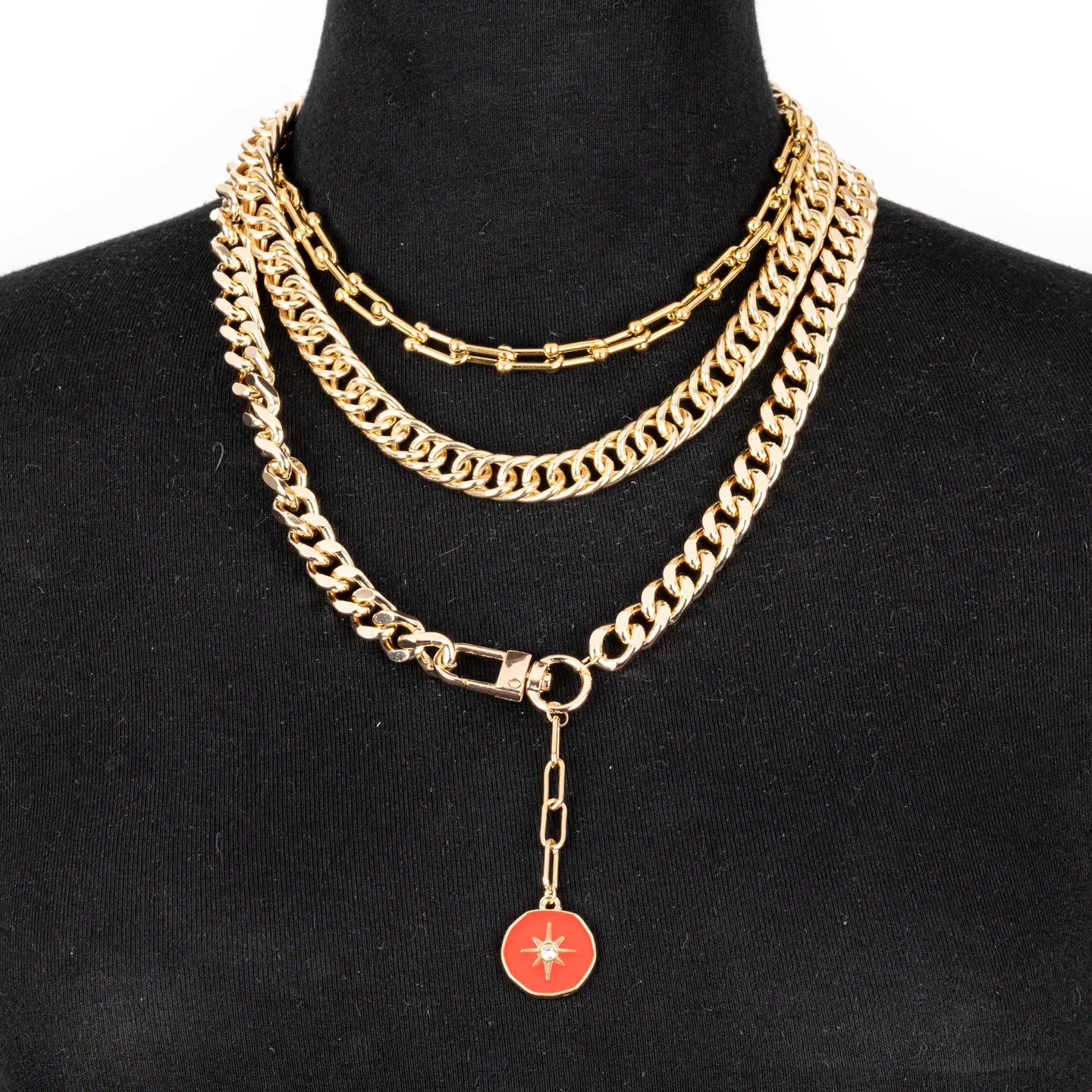 Layered gold chain necklace with multiple chain styles and a red enamel charm drop, displayed on a white background.