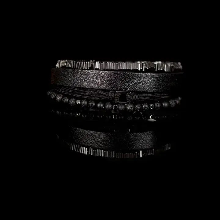 Men’s layered bracelet set featuring a black textured leather cuff, matte black lava stone beads, black braided cord wrap, and gunmetal geometric metal beads with industrial detailing, stacked in a rugged modern luxury design on a white background.