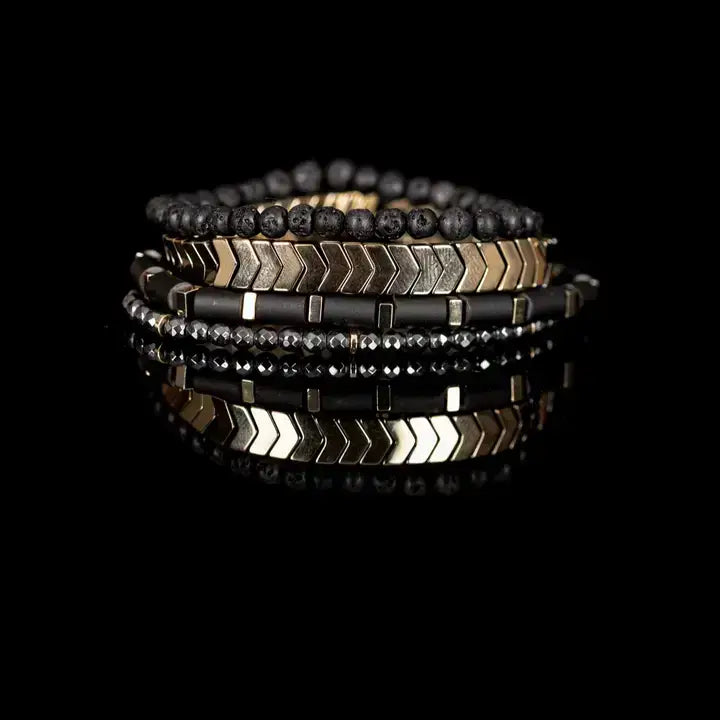 Men’s layered bracelet set featuring matte black lava stone beads, gold-tone chevron metal beads, black cylindrical beads with gold spacers, and faceted hematite beads, stacked in a modern luxury design on a white background.