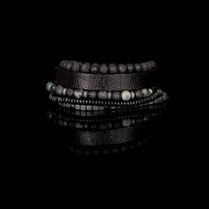Men’s layered bracelet set featuring a black textured leather cuff, matte black lava stone beads, gray marbled stone beads, hematite cube beads, and black heishi spacers, stacked in a rugged contemporary luxury design on a white background.
