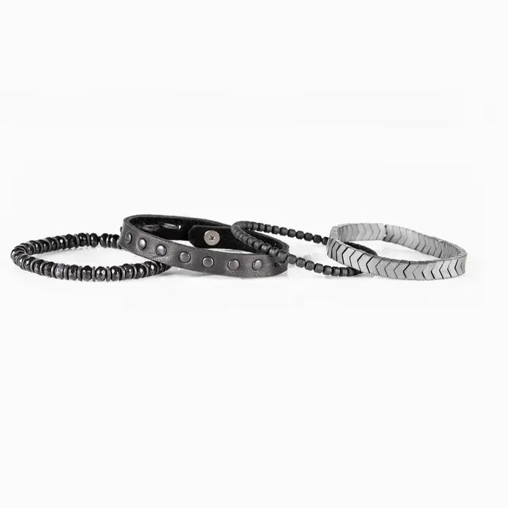 Men’s layered bracelet set featuring a silver-tone chevron stretch bracelet, black leather cuff with gunmetal stud details, matte black bead strand, and faceted black stone beads, stacked in a rugged contemporary luxury design on a white background.