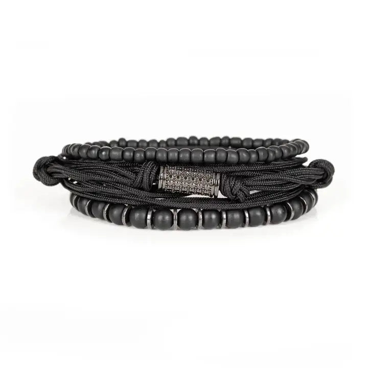 Black layered men’s bracelet set with matte black beads, braided rope wraps, metallic spacer accents, and pavé cylindrical centerpiece on white background.