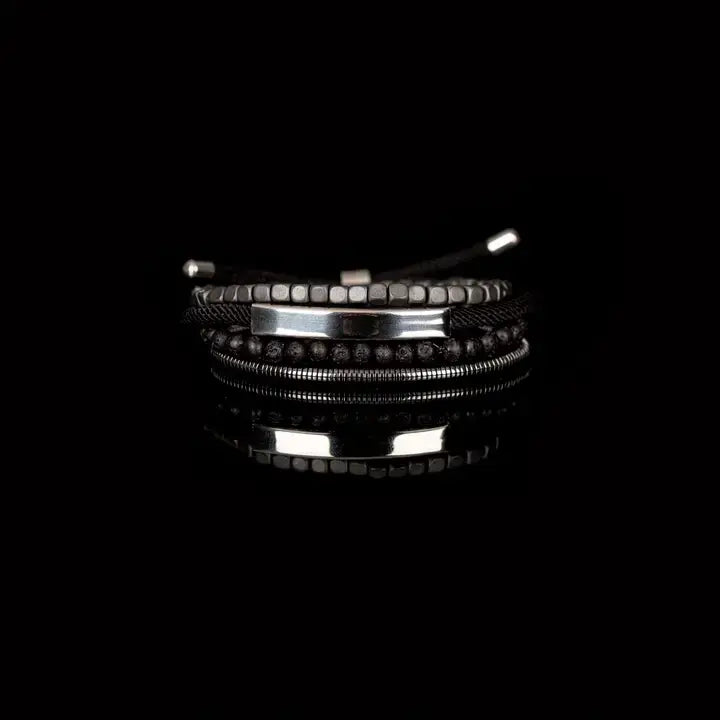 Men’s layered bracelet set featuring a polished silver-tone ID bar centerpiece, matte black lava stone beads, gunmetal cube beads, black braided cord wraps, and silver-tone coil wrap accents, stacked in a sleek industrial luxury design on a white background.