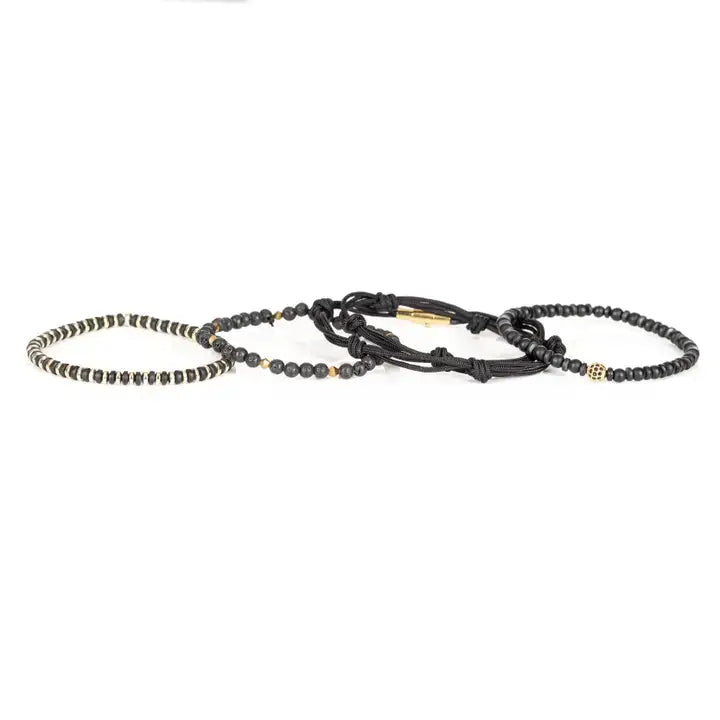 Black and gold layered men’s bracelet set with matte black beads, lava stone accents, gold pavé centerpiece bead, metallic spacers, and braided rope wraps on white background.