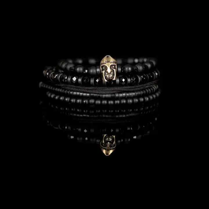 Black multi-layer men’s bracelet featuring matte black beads, faceted onyx stones, metallic tribal mask centerpiece, braided rope layers, and hematite bead strands on a light background.