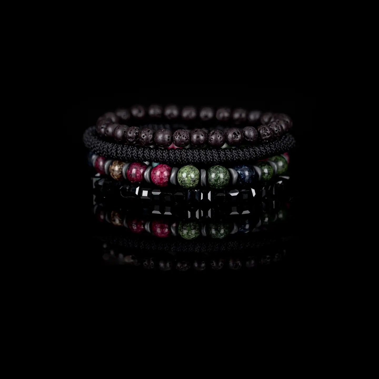 Men's layered bracelet set featuring faceted black crystal cube beads, multicolor natural gemstone beads in green, burgundy, navy, and beige tones, black woven cord detailing, and matte black lava stone beads arranged in a stacked design on a white background.