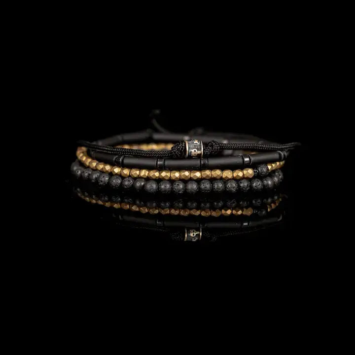 Black and gold multi-layer men’s bracelet featuring matte black tubular beads, faceted gold hematite strands, braided black cord, lava stone beads, and crystal accent detail on a light background.