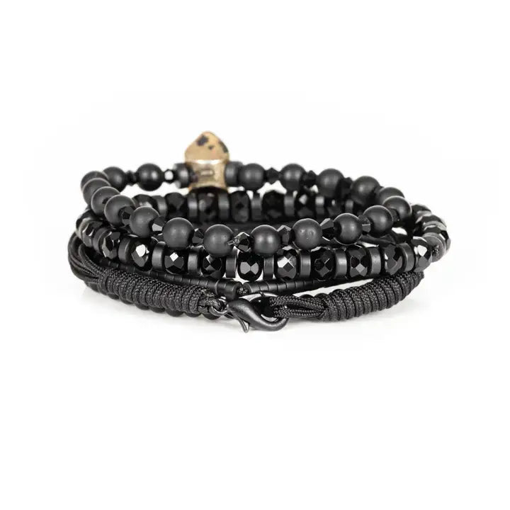 Black multi-layer men’s bracelet featuring matte black beads, faceted onyx stones, metallic tribal mask centerpiece, braided rope layers, and hematite bead strands on a light background.