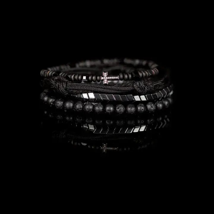 Black layered men’s bracelet set with lava stone beads, pavé cross detail, silver chevron metal strand, braided rope wraps, and woven texture accents on white background.