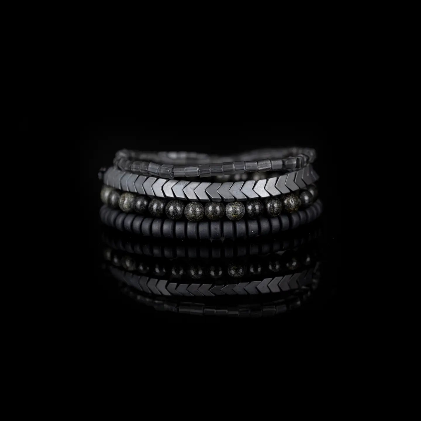 Men’s layered bracelet set featuring gunmetal chevron metal beads, smoky gray translucent beads, black matte heishi beads, and dark gray natural stone beads with subtle metallic shimmer, stacked in a sophisticated monochromatic luxury design on a white background.