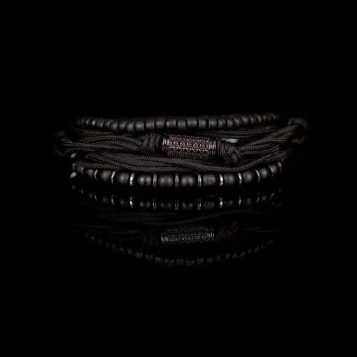 Black layered men’s bracelet set with matte black beads, braided rope wraps, metallic spacer accents, and pavé cylindrical centerpiece on white background.
