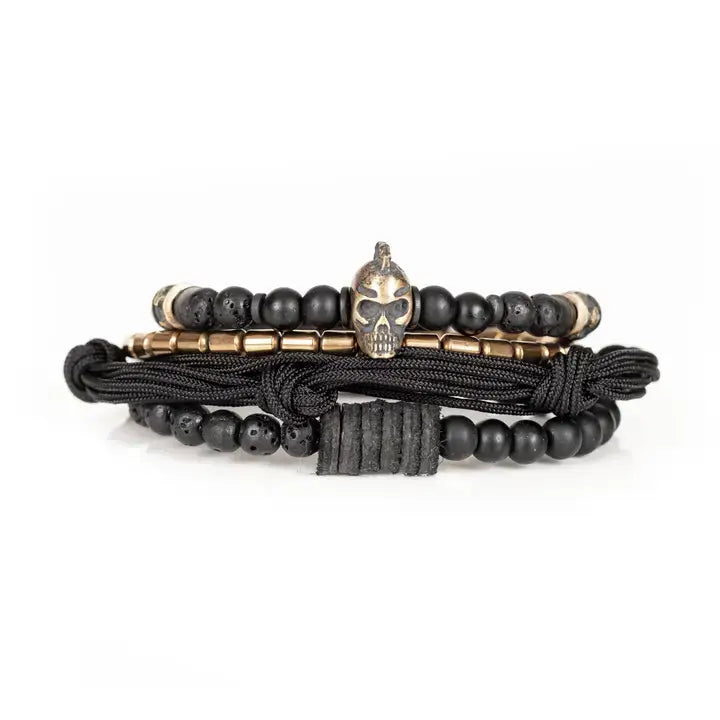 Black layered men’s bracelet set with lava stone beads, metallic skull centerpiece, gold-tone spacer accents, matte black beads, and braided rope wraps on white background.