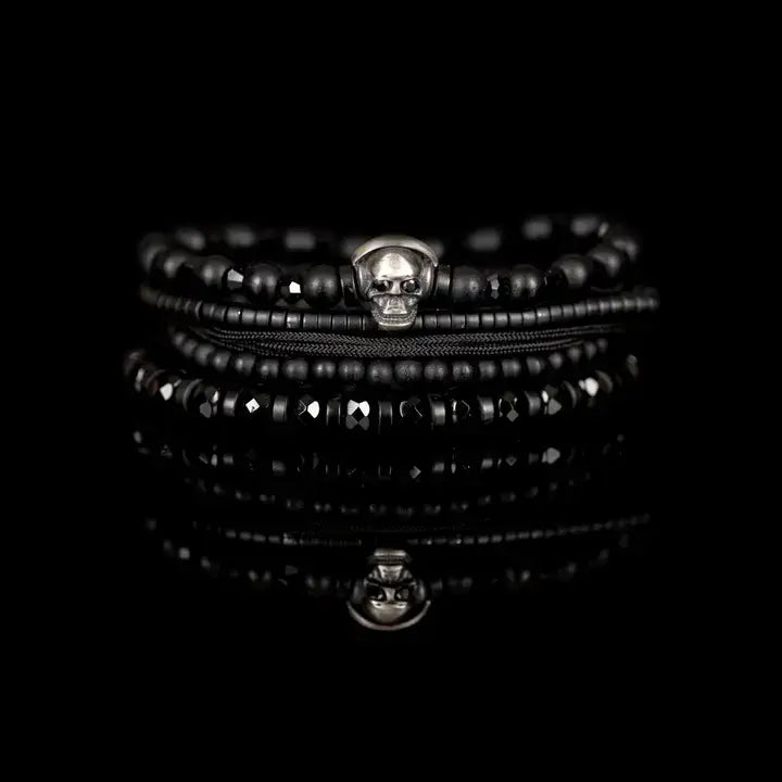 Black layered men’s bracelet set with matte black beads, silver skull centerpiece, faceted crystal accents, and braided rope wraps on white background.