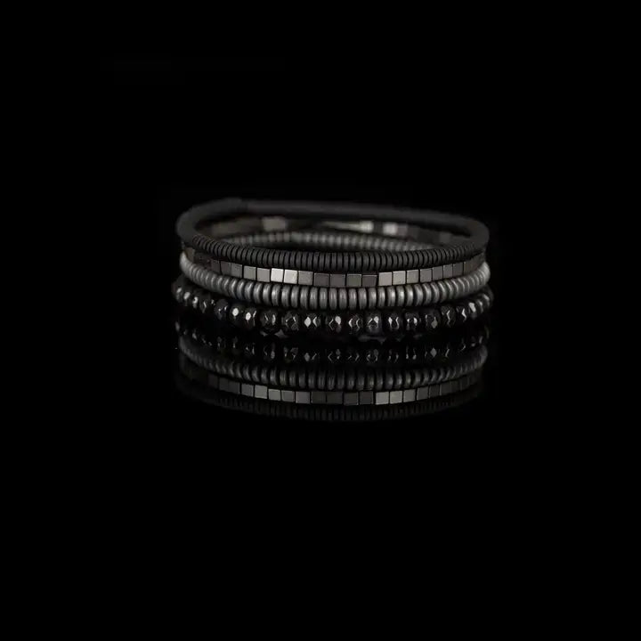 Men's black and gunmetal layered bracelet set featuring black leather, matte black heishi beads, gunmetal cube beads, metallic disc beads, and faceted black crystal beads in a sleek stacked design on a white background.