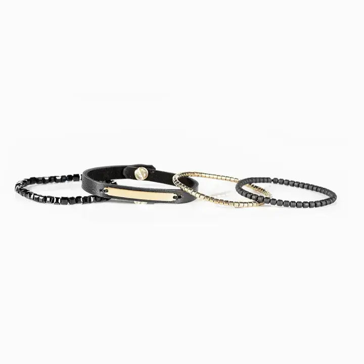 Men’s layered bracelet set featuring a black leather cuff with a gold-tone ID plate, matte black bead strand, gold-tone heishi beads, and faceted black crystal beads, stacked in a sleek modern luxury design on a white background.