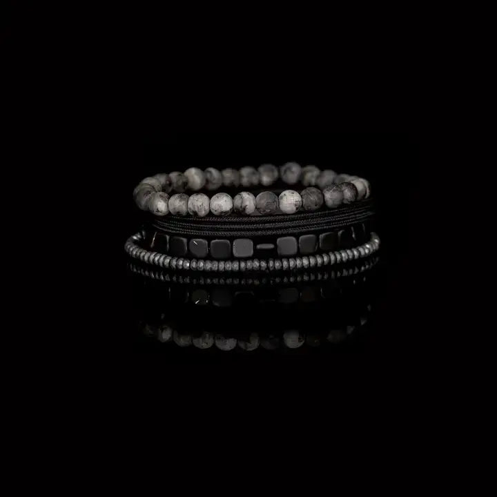 Men’s layered bracelet set featuring gray marbled natural stone beads, polished black hematite cube beads, black braided cord wraps, and silver-gray heishi beads, stacked in a sleek monochromatic luxury design on a white background