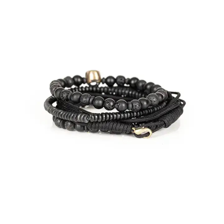 Black layered men’s bracelet set with matte beads, lava stone accents, braided rope wraps, and metallic skull centerpiece on white background.