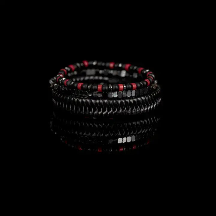 Black multi-layer men’s bracelet featuring glossy black disc beads, metallic hematite cube beads, matte black accents, and red bead details with adjustable cord closure on a light background.