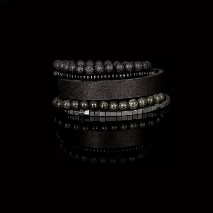 Men’s layered bracelet set featuring a black leather cuff, matte black lava stone beads, dark gray natural stone beads, black heishi spacers, faceted black crystal beads, and hematite cube beads, stacked in a sophisticated monochromatic luxury design on a white background.