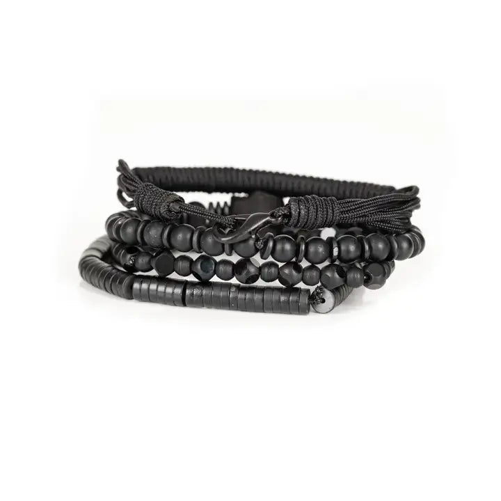 Men's black layered bracelet set featuring braided cord, matte black beads, faceted black crystal beads, a carved skull centerpiece, and black heishi disc beads, arranged in a stacked design on a white background.