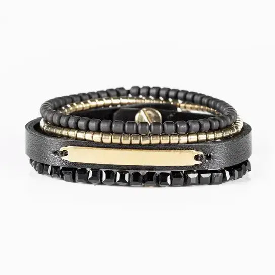 Men’s layered bracelet set featuring a black leather cuff with a gold-tone ID plate, matte black bead strand, gold-tone heishi beads, and faceted black crystal beads, stacked in a sleek modern luxury design on a white background.