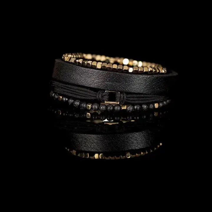 Men’s layered bracelet set featuring a black textured leather cuff, gold-tone faceted metal bead bracelet, black braided cord wrap, matte black lava stone beads, and gold-tone accent beads, stacked in a refined modern luxury design on a white background.