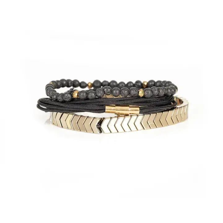 Black and gold layered men’s bracelet with lava stone beads, braided rope wraps, gold chevron metal bar, and metallic accents on white background.