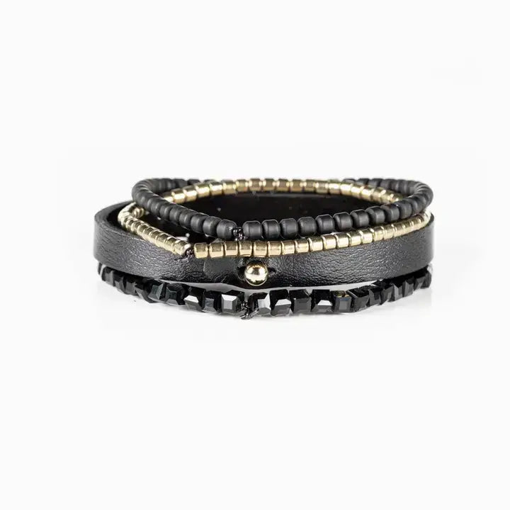Men’s layered bracelet set featuring a black leather cuff with a gold-tone ID plate, matte black bead strand, gold-tone heishi beads, and faceted black crystal beads, stacked in a sleek modern luxury design on a white background.