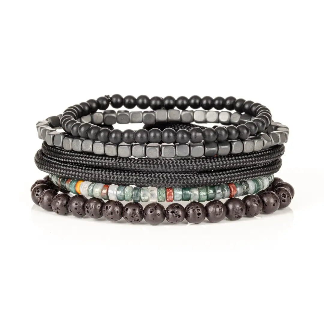 Men’s layered bracelet set featuring matte black bead strands, gunmetal cube beads, black braided cord wraps, dark lava stone beads, and green natural stone accent beads with subtle earth-tone details, stacked in a rugged contemporary luxury design on a white background.