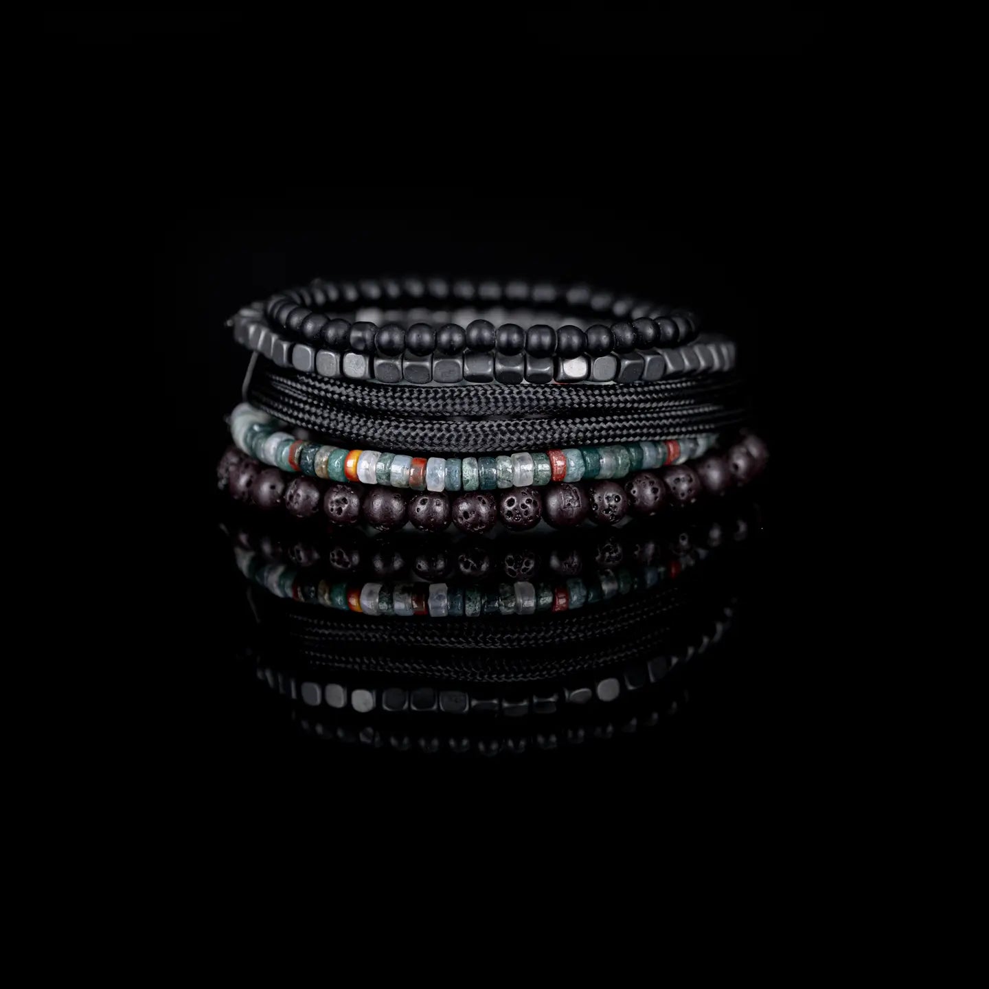 Men’s layered bracelet set featuring matte black bead strands, gunmetal cube beads, black braided cord wraps, dark lava stone beads, and green natural stone accent beads with subtle earth-tone details, stacked in a rugged contemporary luxury design on a white background.