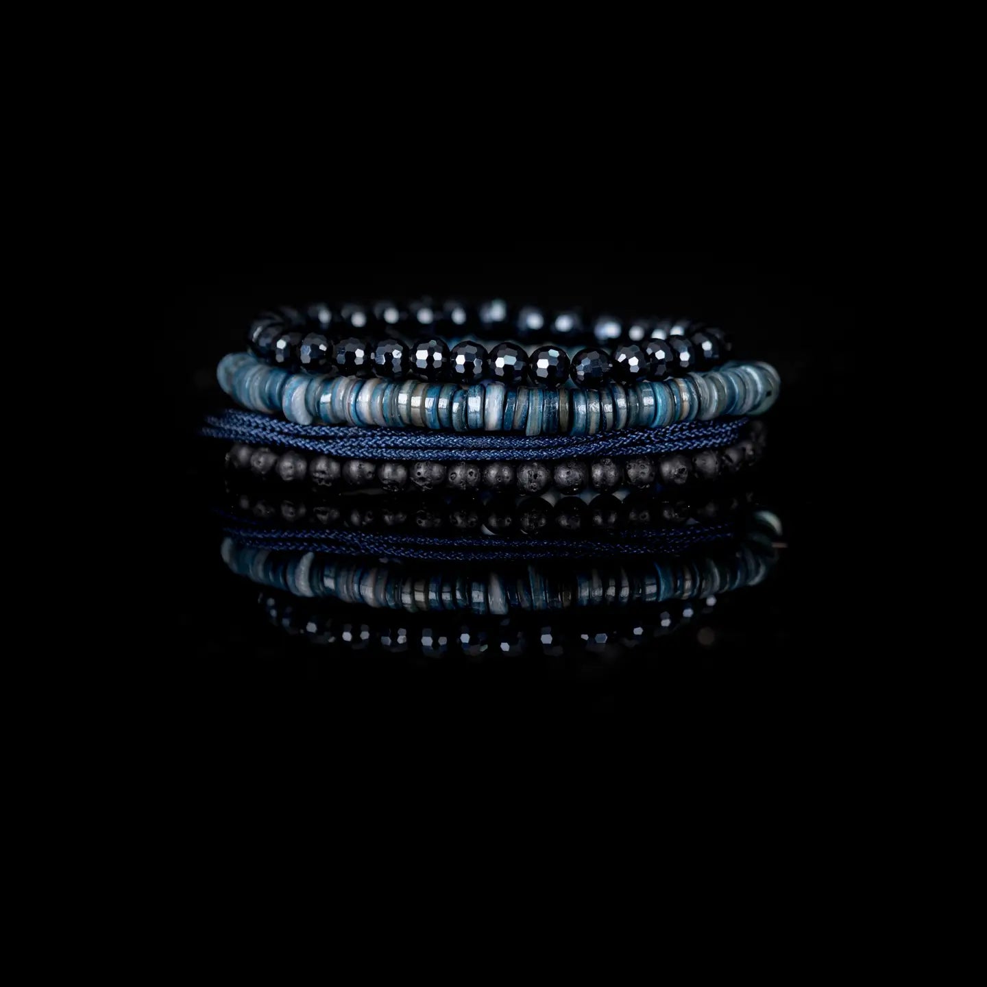 Men's layered bracelet set featuring faceted black crystal beads, blue heishi stone beads, navy woven cord, and matte black lava stone beads arranged in a stacked design on a white background.