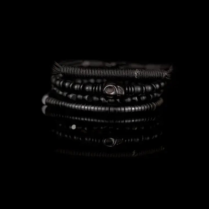 Men's black layered bracelet set featuring braided cord, matte black beads, faceted black crystal beads, a carved skull centerpiece, and black heishi disc beads, arranged in a stacked design on a white background.