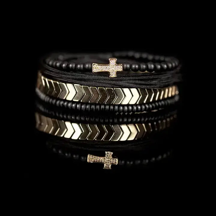 Black multi-layer men’s bracelet set featuring matte onyx beads, braided black rope, crystal cross centerpiece, gold chevron metal bar, and layered metallic accents on a light background.