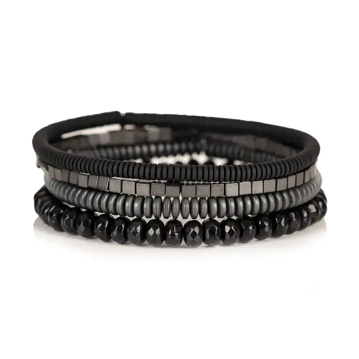 Men's black and gunmetal layered bracelet set featuring black leather, matte black heishi beads, gunmetal cube beads, metallic disc beads, and faceted black crystal beads in a sleek stacked design on a white background.