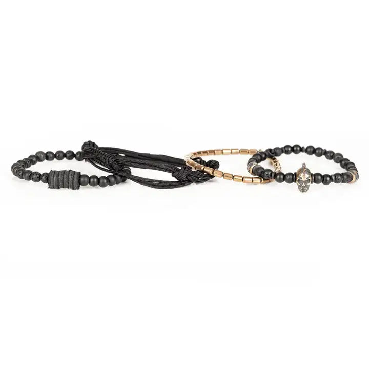 Black layered men’s bracelet set with lava stone beads, metallic skull centerpiece, gold-tone spacer accents, matte black beads, and braided rope wraps on white background.