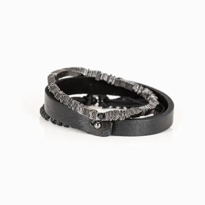 Men’s layered bracelet set featuring a black textured leather cuff, matte black lava stone beads, black braided cord wrap, and gunmetal geometric metal beads with industrial detailing, stacked in a rugged modern luxury design on a white background.