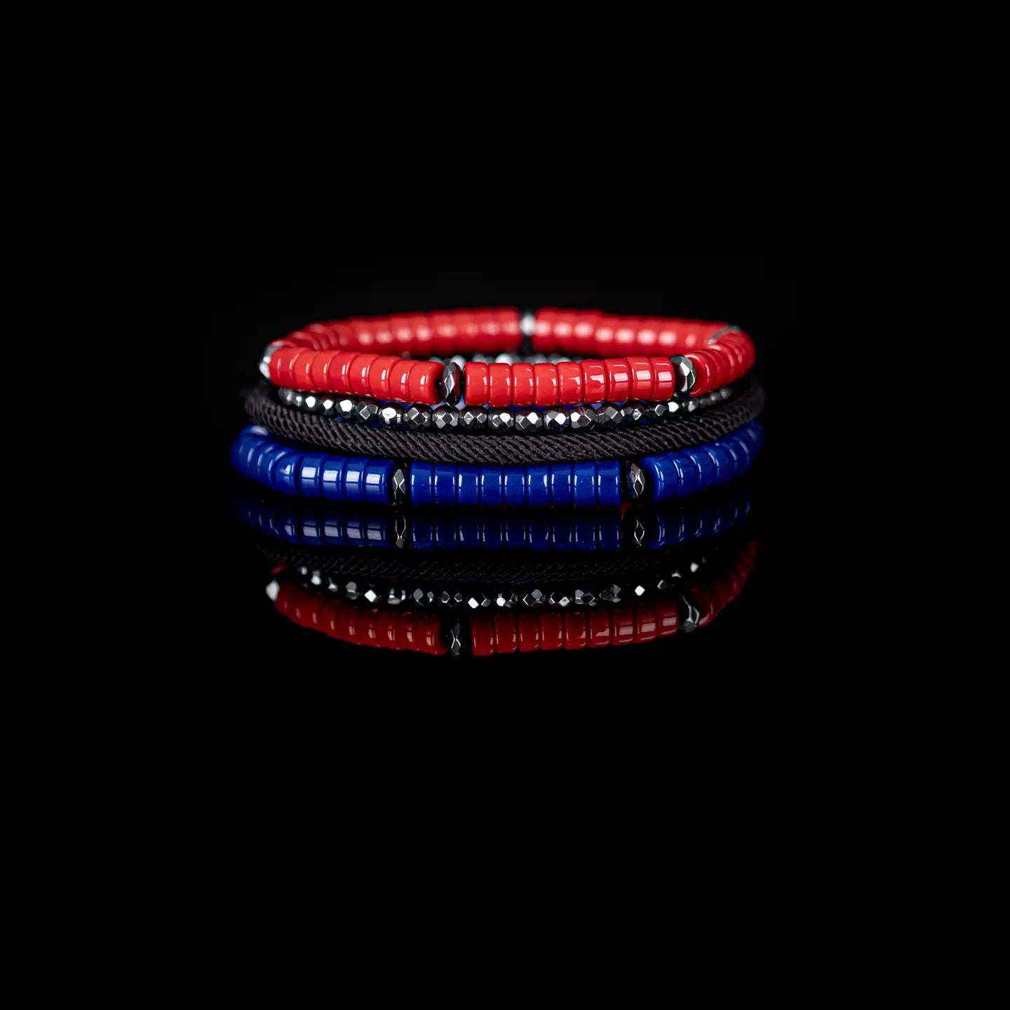 Men’s layered bracelet set featuring vibrant red and royal blue heishi beads, black braided cord, faceted hematite crystal beads, and gunmetal spacer accents, stacked in a bold contemporary luxury design on a white background.