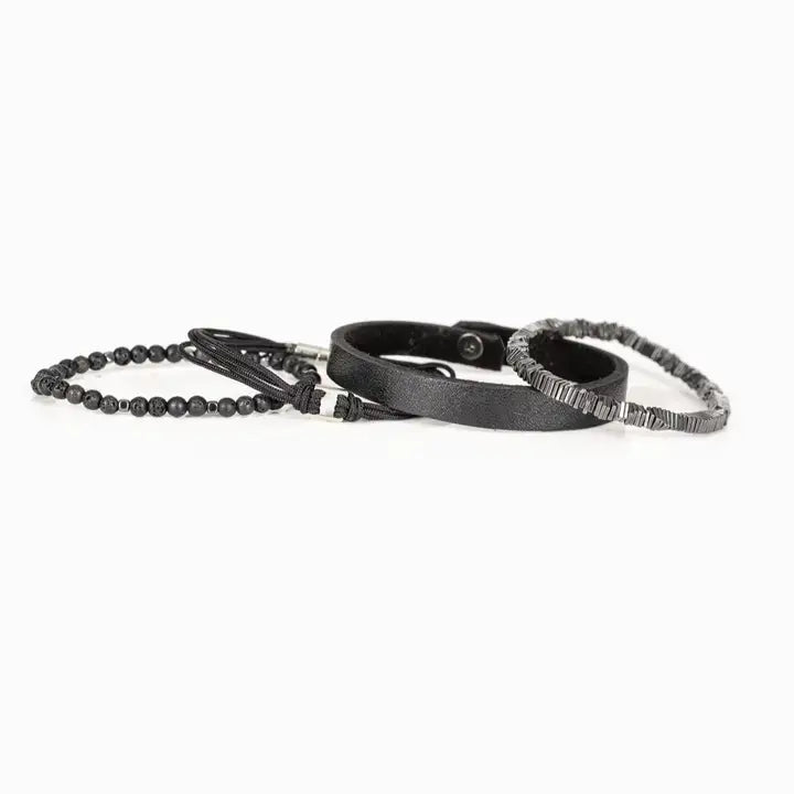 Men’s layered bracelet set featuring a black textured leather cuff, matte black lava stone beads, black braided cord wrap, and gunmetal geometric metal beads with industrial detailing, stacked in a rugged modern luxury design on a white background.