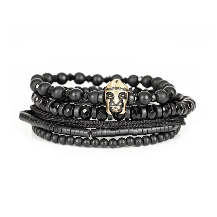 Black multi-layer men’s bracelet featuring matte black beads, faceted onyx stones, metallic tribal mask centerpiece, braided rope layers, and hematite bead strands on a light background.