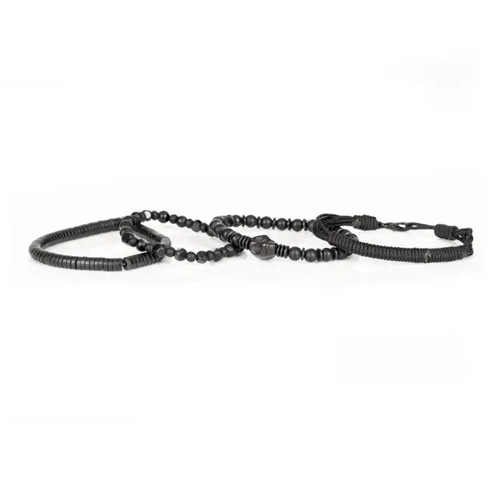 Men's black layered bracelet set featuring braided cord, matte black beads, faceted black crystal beads, a carved skull centerpiece, and black heishi disc beads, arranged in a stacked design on a white background.