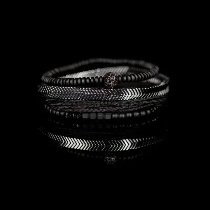 Black multi-layer men’s bracelet featuring silver chevron metal bar, hematite bead strands, crystal accent bead, faceted black stones, and braided black rope layers on a light background.